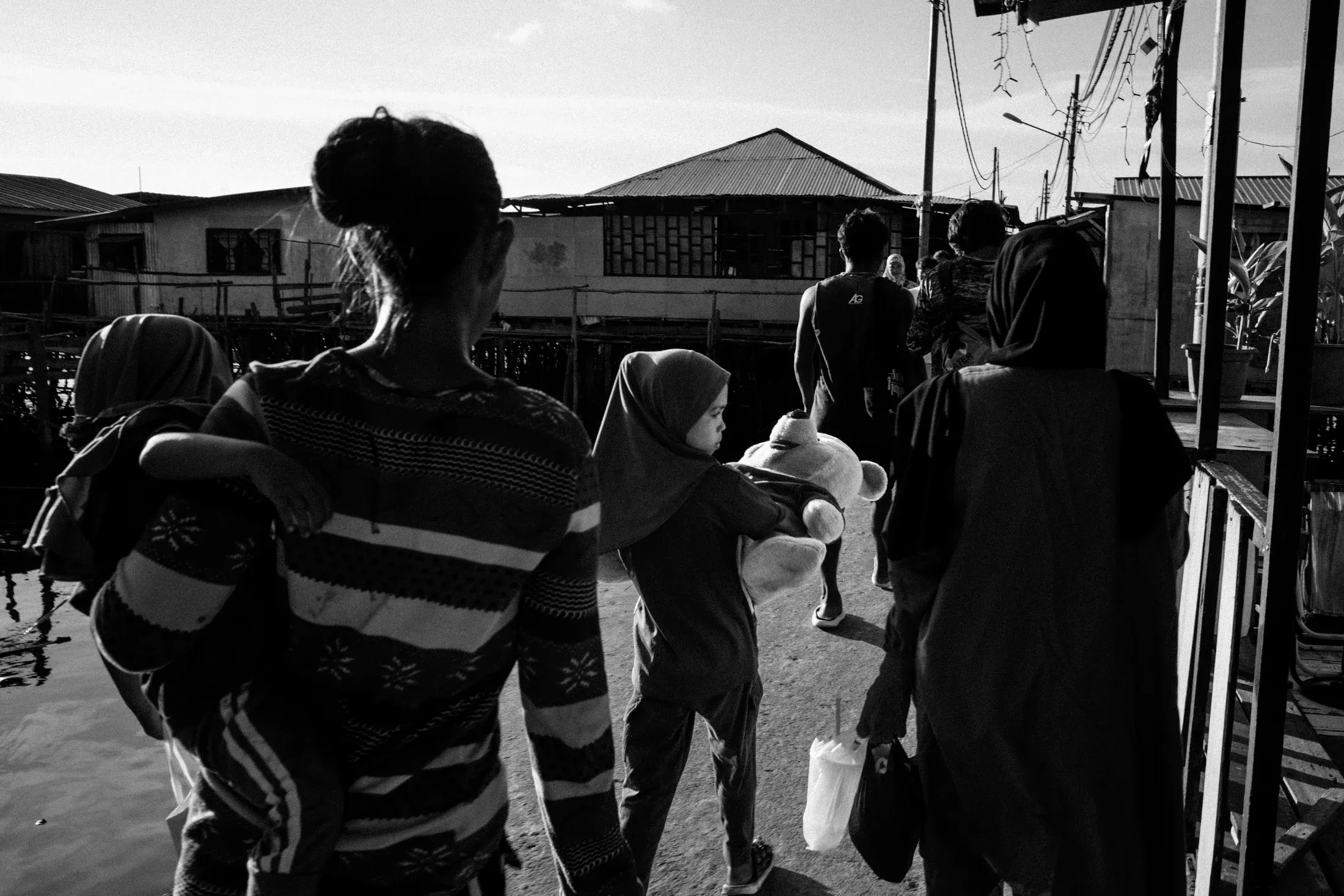 Black and white view from behind of residents moving through a narrow stilt-house walkway.
