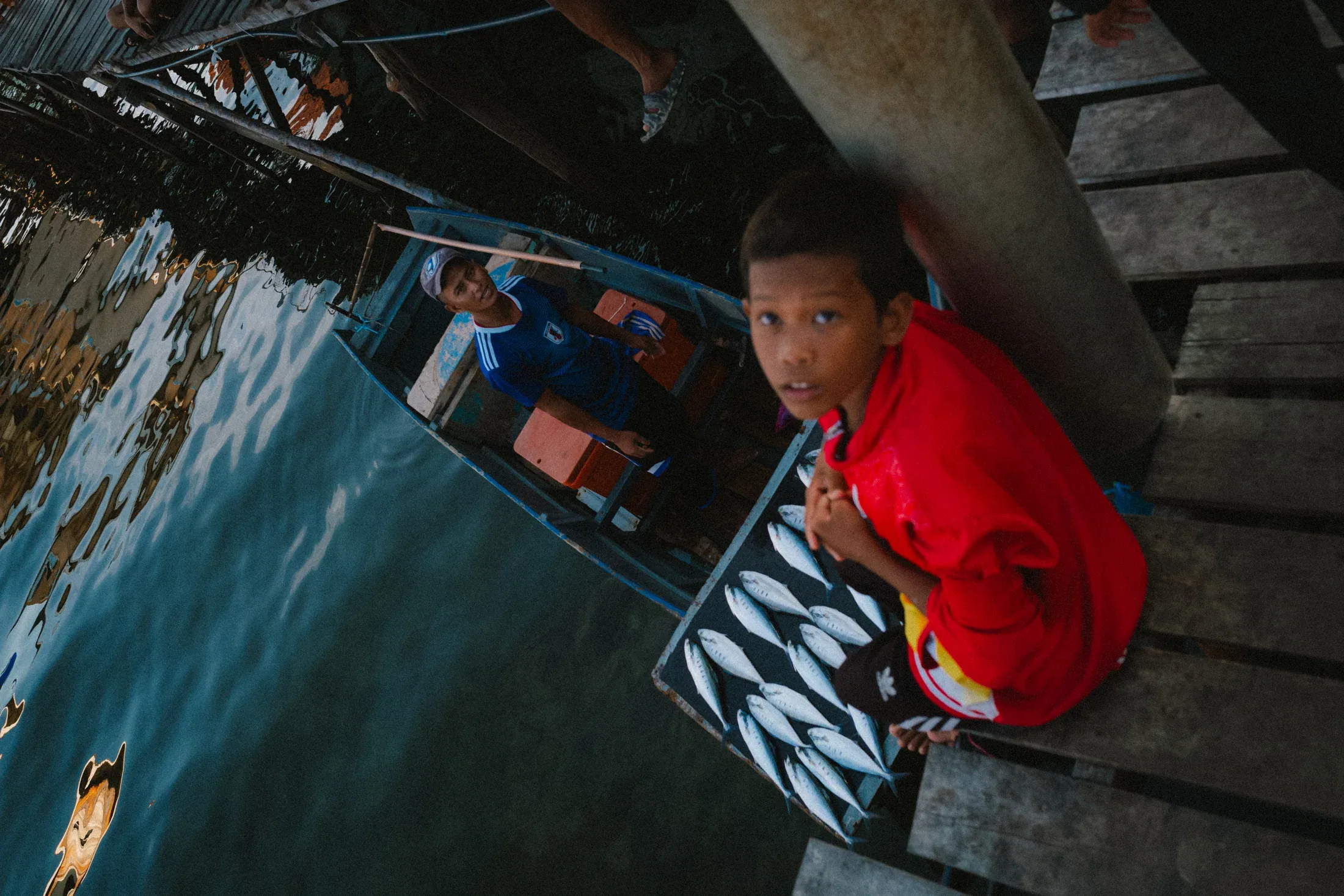 Boy in a red shirt looking up from a wooden walkway above dark water.