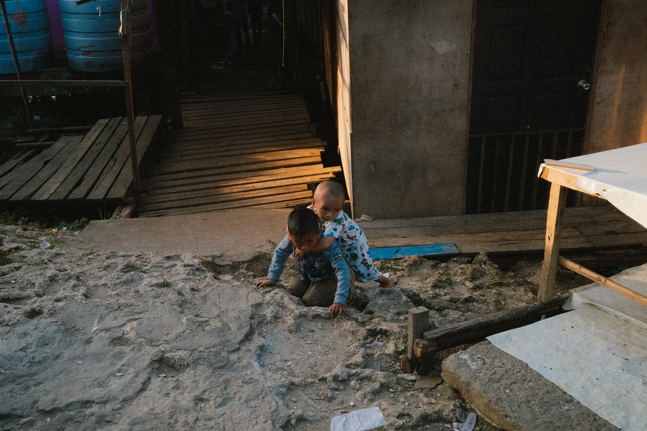 Small child sitting alone in sand at the shaded entrance of a wooden house.