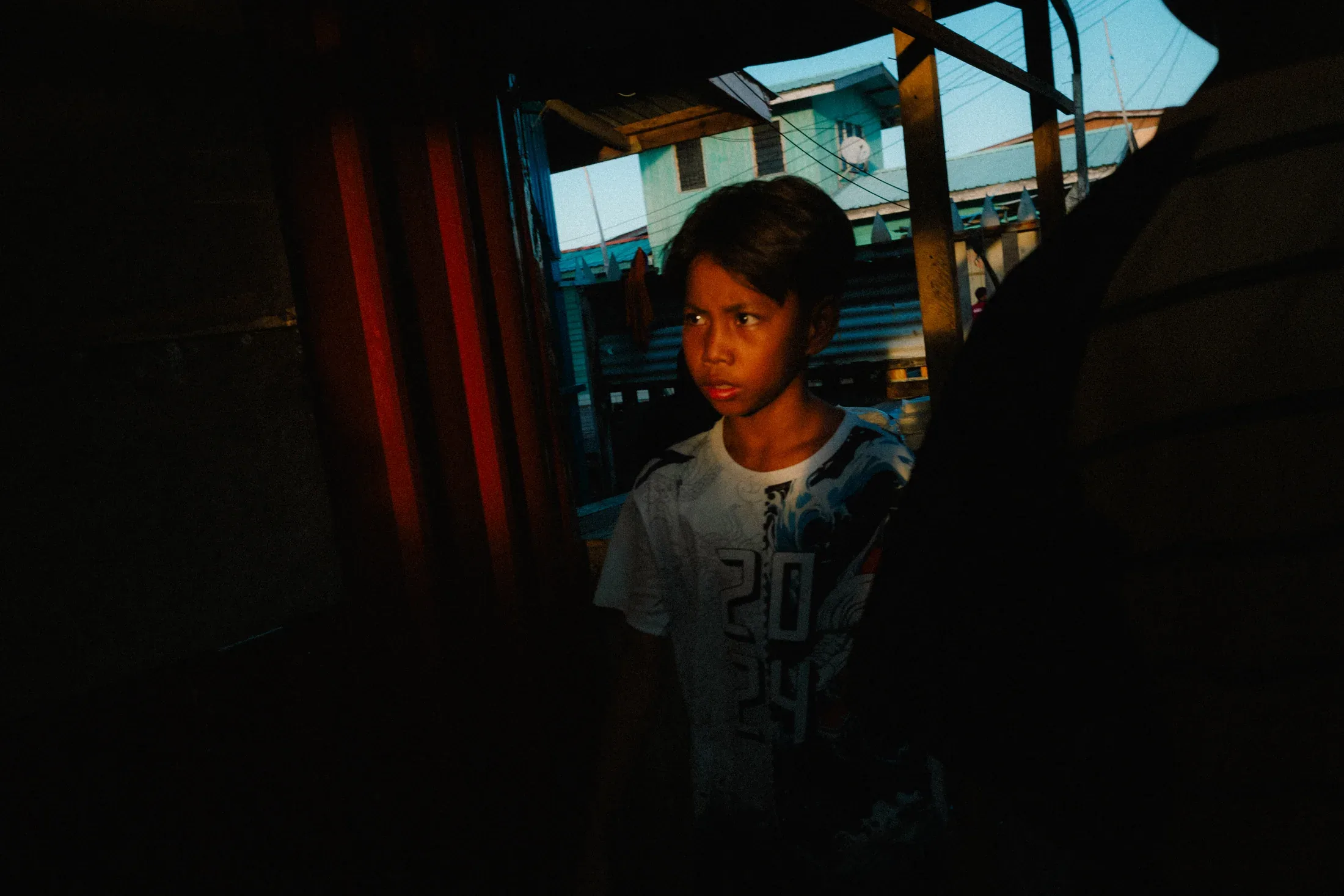 Girl standing in a dark doorway framed by blue daylight and deep wooden shadows.