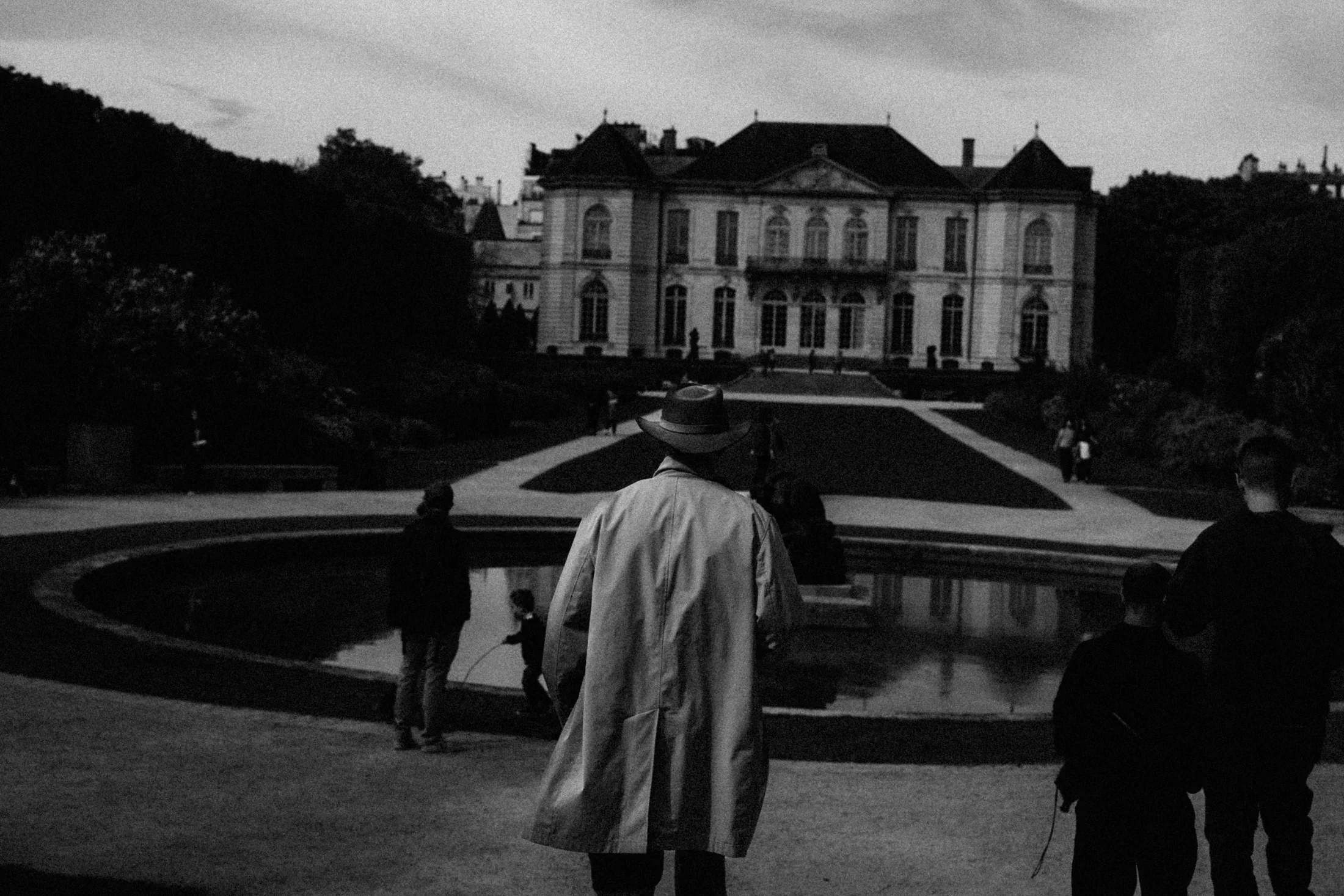 Man in a light coat walking toward a formal garden and grand house in black and white.